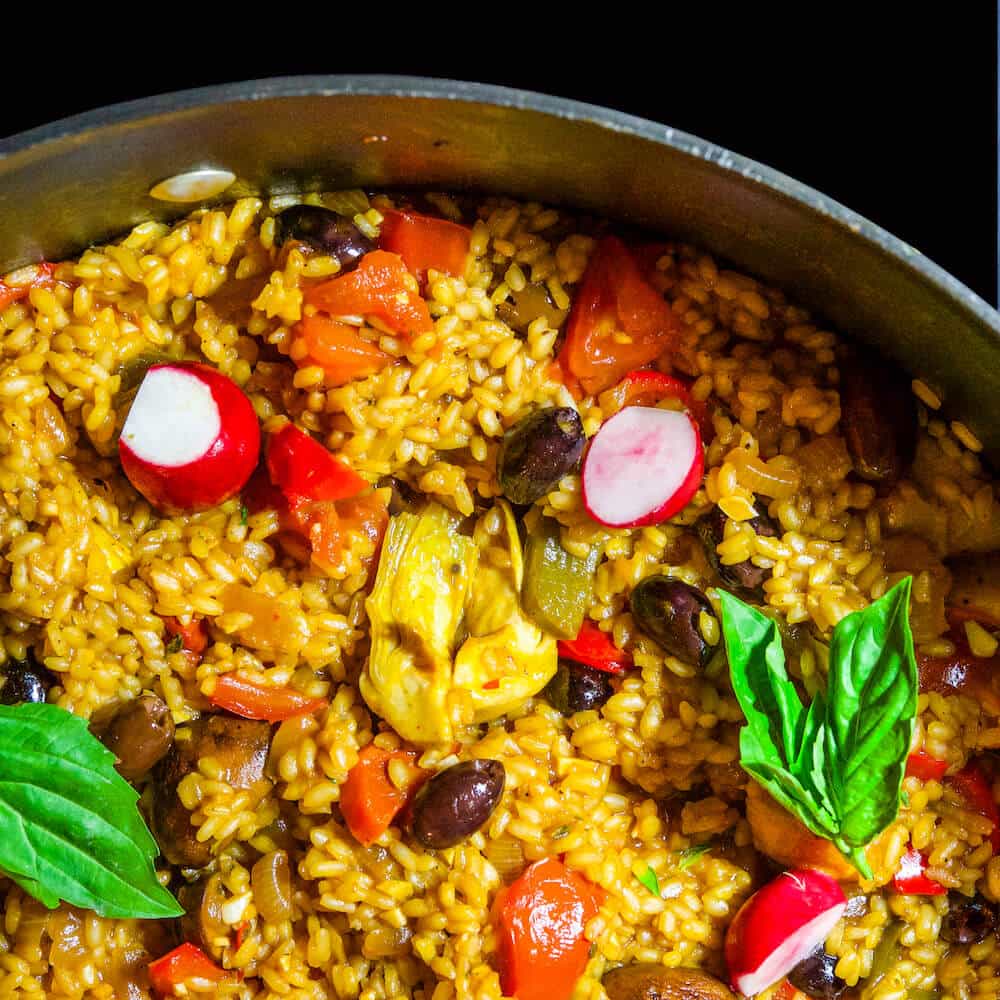 A close-up of a pan containing yellow rice mixed with vegetables, including radish slices, olives, artichokes, tomatoes, and basil leaves.