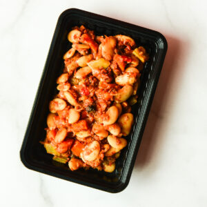A black plastic container of Plaki, cooked lima beans in tomato-based sauce, sits on a white surface.