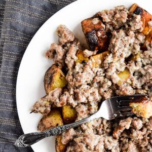 A white plate of Plaki—roasted potato chunks topped with ground sausage and gravy—served with a fork.