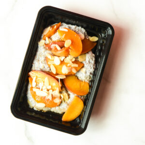 Dukes Of Peaches chia pudding in a black container, topped with sliced peaches and slivered almonds on a white surface.