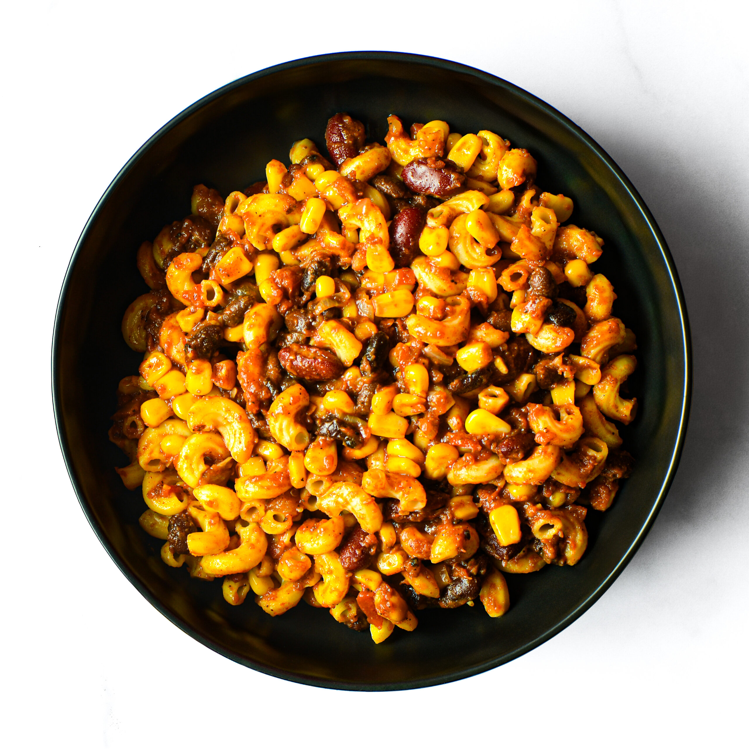 The Redneck Riviera: a black bowl filled with macaroni, corn, kidney beans, ground beef, and tomato sauce, shot from above on a white background.