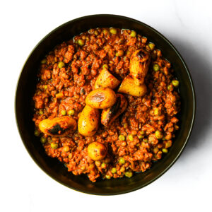 The Redneck Riviera: Minced meat curry with green peas, topped with roasted baby potatoes, served on a white surface.