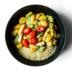 The Redneck Riviera: A black bowl with cooked couscous, sautéed gnocchi in creamy spinach sauce, cherry tomatoes, and sliced white veggies.