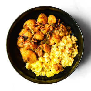 The Redneck Riviera: A black bowl with yellow scrambled eggs and seasoned roasted potatoes, peppers, and onions on a white background.