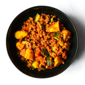 A top view of The Redneck Riviera, a black bowl with lentil and vegetable curry featuring potatoes, zucchini, and tomatoes on a white background.
