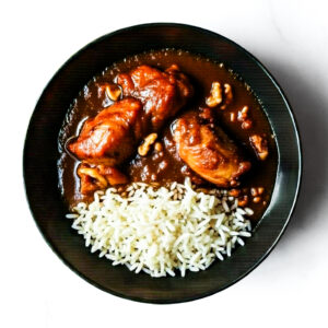 The Redneck Riviera: Chicken in brown sauce over white rice, garnished with nuts, served on a black plate and set on a white surface.