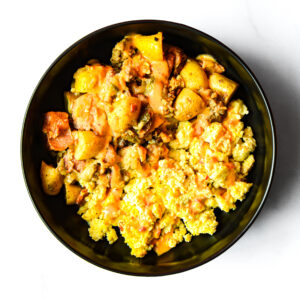The Redneck Riviera: scrambled eggs with sautéed potatoes, onions, and melted cheese in a black bowl, viewed from above on a white surface.