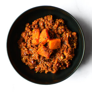 A black bowl of The Redneck Riviera chili, featuring ground meat, beans, and orange squash chunks, seen from above on a white background.