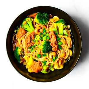 The Redneck Riviera: a black bowl with noodles, broccoli, cashews, green onions, and red sauce on a white surface.