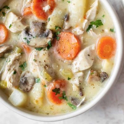 A close-up of a hearty chicken stew filled with chunks of chicken, sliced carrots, mushrooms, potatoes, and celery in a creamy broth. Garnished with chopped parsley.