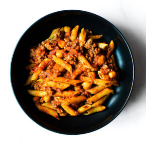 A black bowl of Mac To The Face—penne pasta, ground meat, chickpeas, and tomato sauce—seen from above on a white surface.