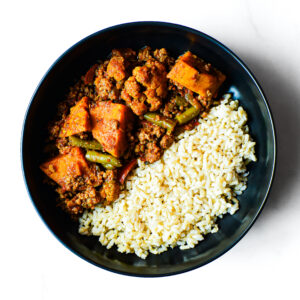 A black bowl with Mac To The Face: cooked white rice topped with savory ground meat, sweet potato, and green bean stew.