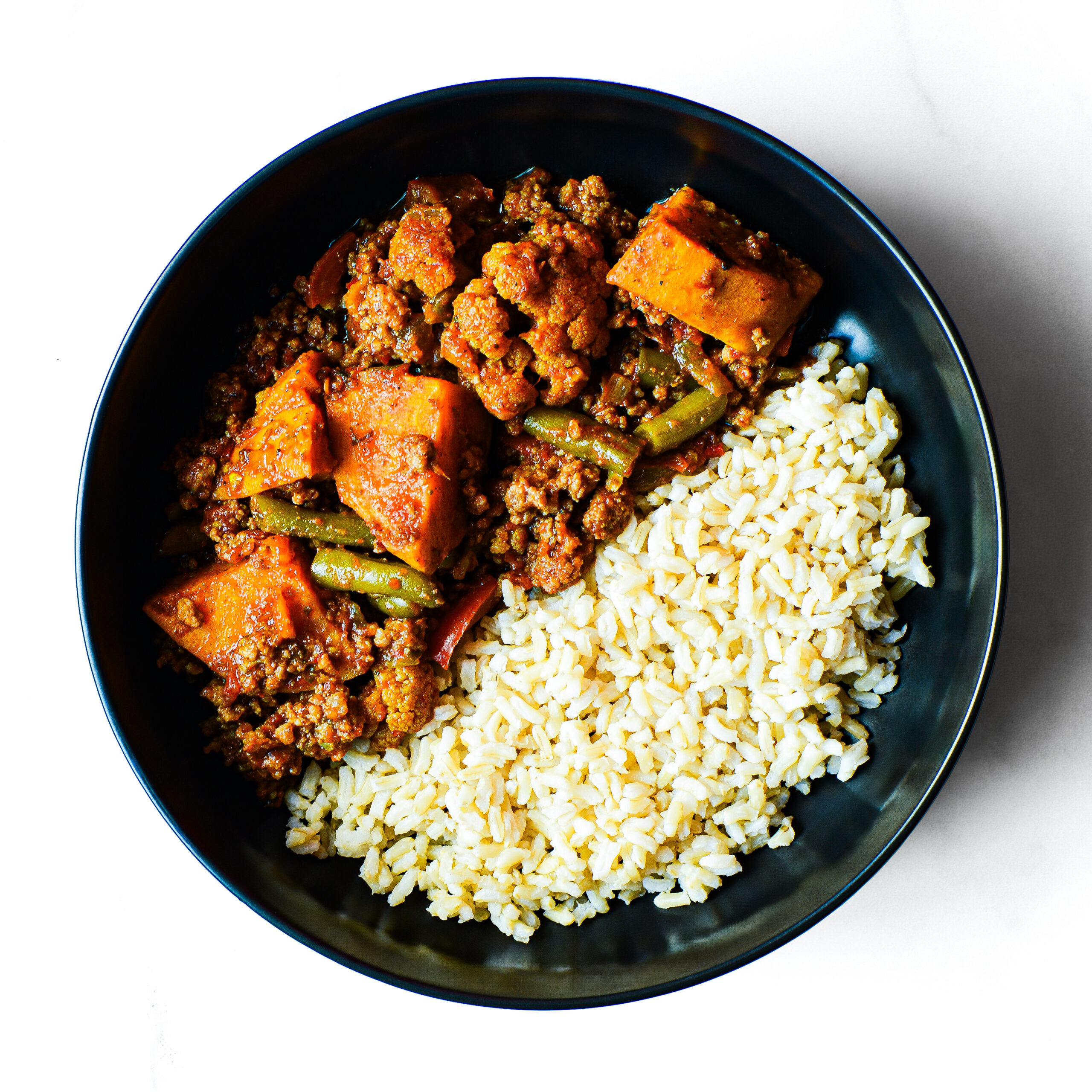 A black bowl with Mac To The Face: cooked white rice topped with savory ground meat, sweet potato, and green bean stew.