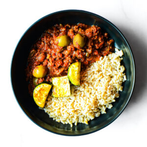 A black bowl of Mac To The Face—white rice, ground meat in tomato sauce, green olives, and cooked zucchini—on a white surface.