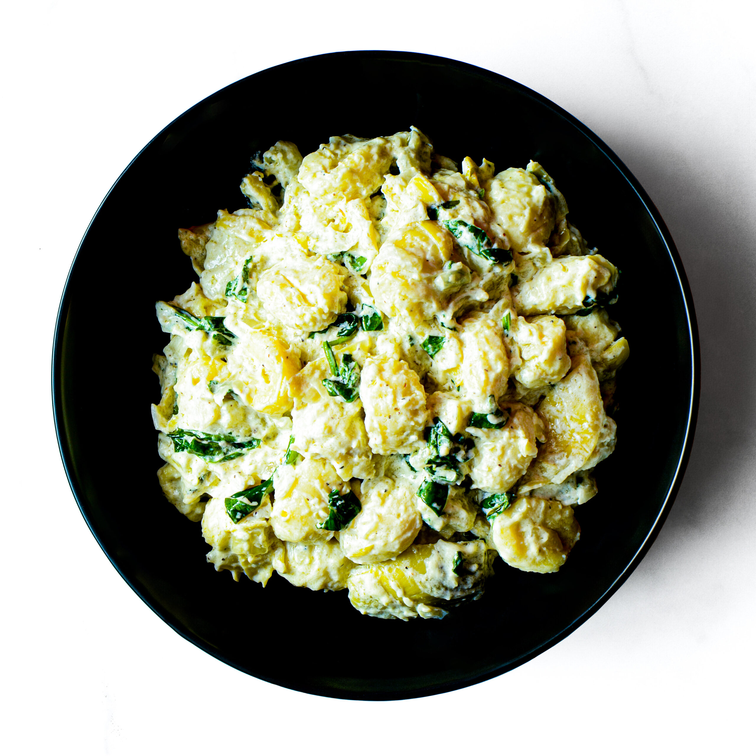 A black bowl of Mac To The Face—creamy pasta with wilted spinach—seen from above on a white background.