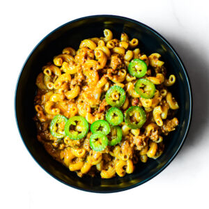 A black bowl of Mac To The Face: macaroni pasta with ground meat, sauce, and sliced green jalapeño peppers on top.