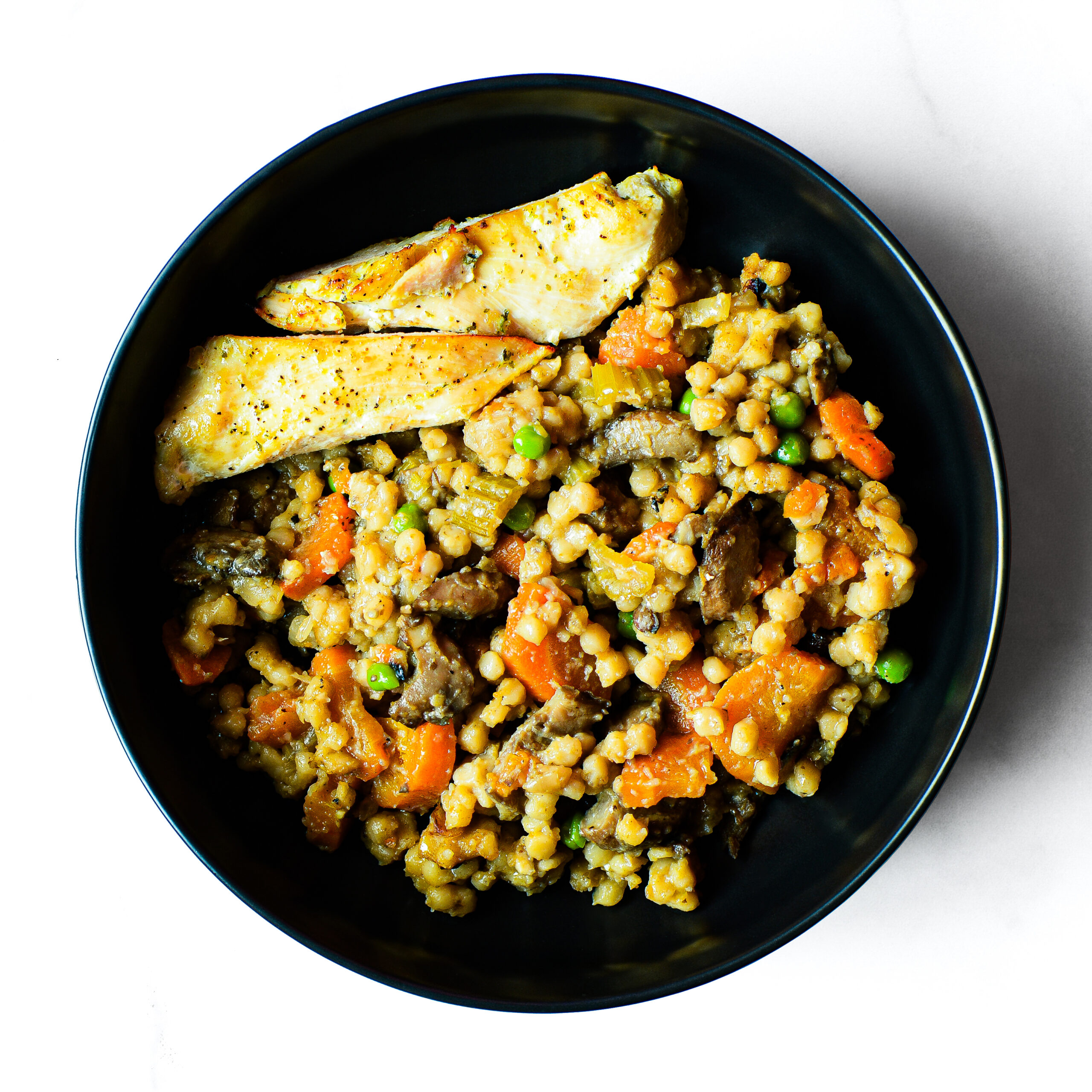 The “Mac To The Face” features a black bowl with barley, vegetables, and sliced chicken, photographed from above on a white background.