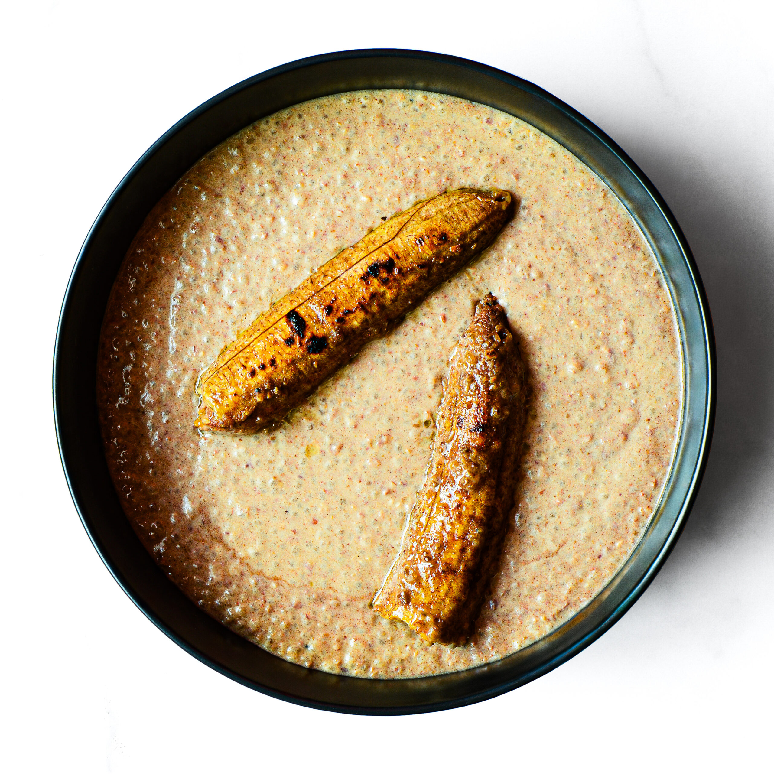 A bowl of Mac To The Face porridge with two pieces of grilled plantain on top, viewed from above on a white background.