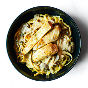 A black bowl of Mac To The Face—creamy pasta with sliced grilled chicken breast and mushrooms—on a white background.