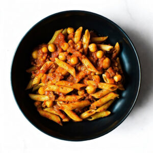 A black bowl of Mac To The Face penne pasta with tomato sauce and chickpeas, photographed from above on a white background.
