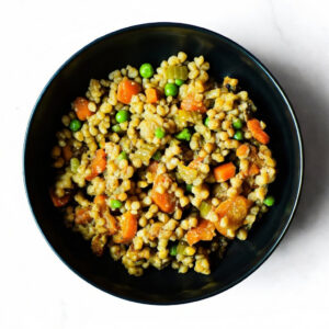 A black bowl of Mac To The Face, a mixed vegetable and pearl couscous salad with carrots, peas, and celery, on a white background.