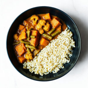 A black bowl of Mac To The Face, a brown rice and vegetable curry with sweet potatoes, green beans, and cauliflower, viewed from above.