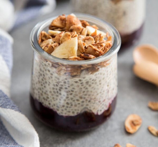 A glass jar filled with chia pudding layered over dark berry compote and topped with granola and banana slices. A striped cloth and a wooden spoon are in the background on a gray surface.