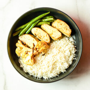 Cluck Norris: A black bowl with white rice, sliced chicken breast, roasted cauliflower, and green beans on light marble.