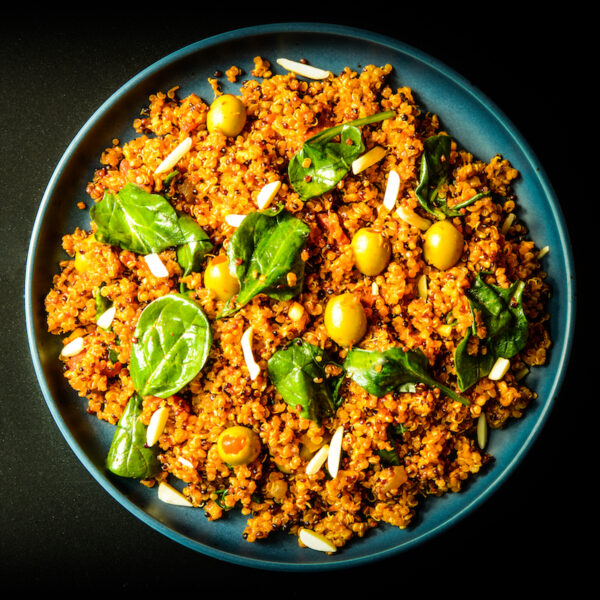 A blue plate filled with a colorful quinoa salad, featuring spinach leaves, green olives, and sliced almonds, all mixed in a vibrant orange seasoning, set against a dark background.