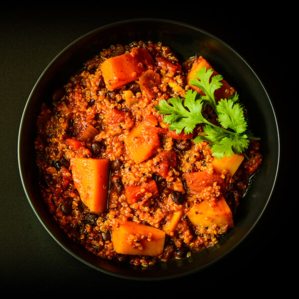 A bowl of quinoa salad with chunks of sweet potatoes, black beans, and tomatoes, garnished with fresh cilantro, served in a dark bowl against a black background.