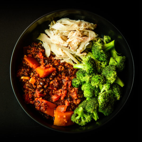 A bowl with shredded chicken, cooked broccoli, and a quinoa mix with black beans, tomatoes, and bell peppers on a dark background.