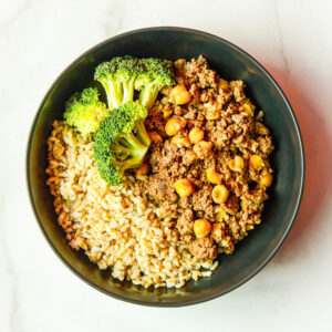 A black bowl with Cluck Norris ground meat, cooked brown rice, chickpeas, and steamed broccoli florets on a white surface.