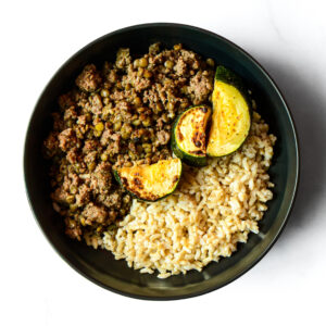 A black bowl of Moo You Didn't! with cooked ground meat, peas, roasted zucchini slices, and brown rice on a white surface.