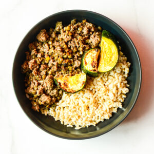 A black bowl of Cluck Norris: ground meat with peas, two slices of cooked zucchini, and brown rice on a white surface.
