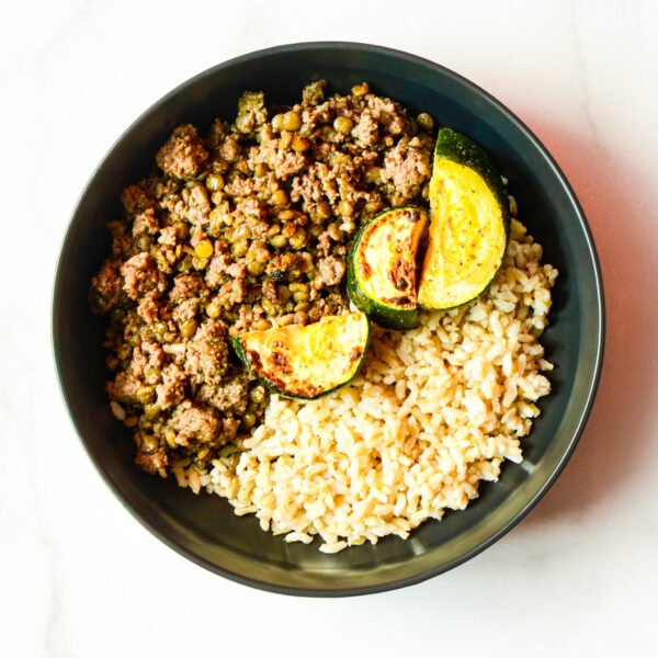 A black bowl of Cluck Norris: ground meat with peas, two slices of cooked zucchini, and brown rice on a white surface.