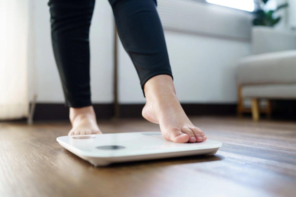 A person steps onto a digital bathroom scale on a wooden floor, with light coming in from a nearby window.
