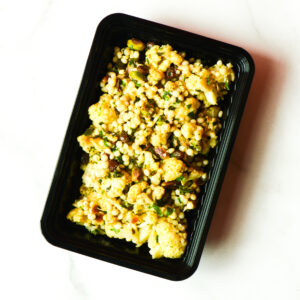 Cluck The Bean in a black container: cauliflower and couscous salad with herbs, nuts, and raisins on a white surface.
