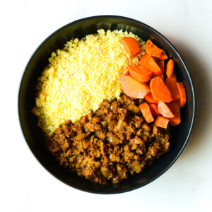 A black bowl of Cluck The Bean: couscous, cooked carrots, and spiced ground meat with onions, each in separate sections.