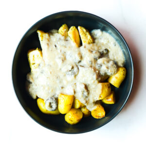 A black bowl of Cluck The Bean topped with creamy mushroom sauce and roasted yellow potatoes, viewed from above on a white surface.