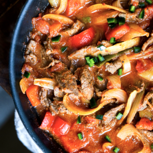 Close-up of BiSteak Rancheros - WL in a pan, with sliced beef, red bell peppers, onions, and green onions in a spicy sauce.