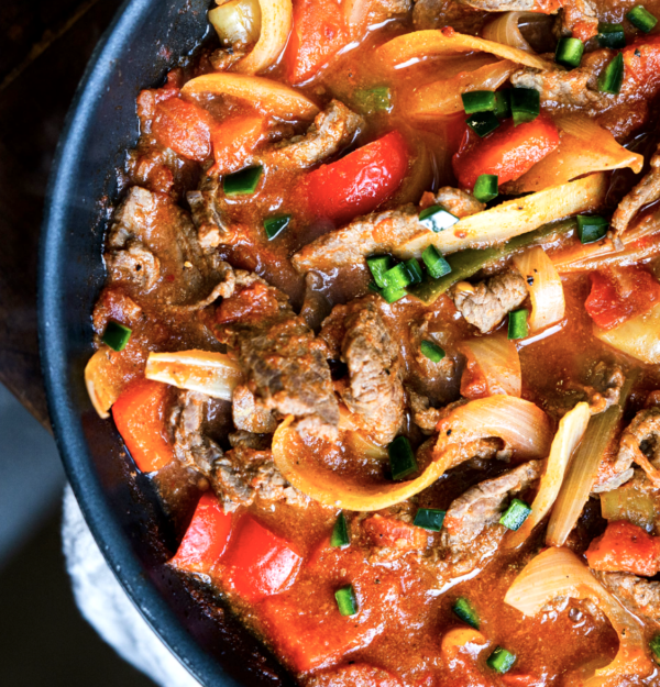 Close-up of BiSteak Rancheros - WL in a pan, with sliced beef, red bell peppers, onions, and green onions in a spicy sauce.