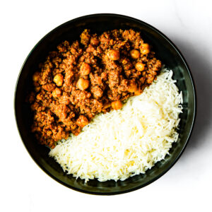 A black bowl of white rice and ground beef-chickpea curry from Operation Cajun Fit, placed side by side on a white surface.