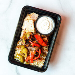 The Mean Mexi-Machine: a black meal prep container with grilled chicken, roasted bell peppers, rice, and white sauce on white marble.