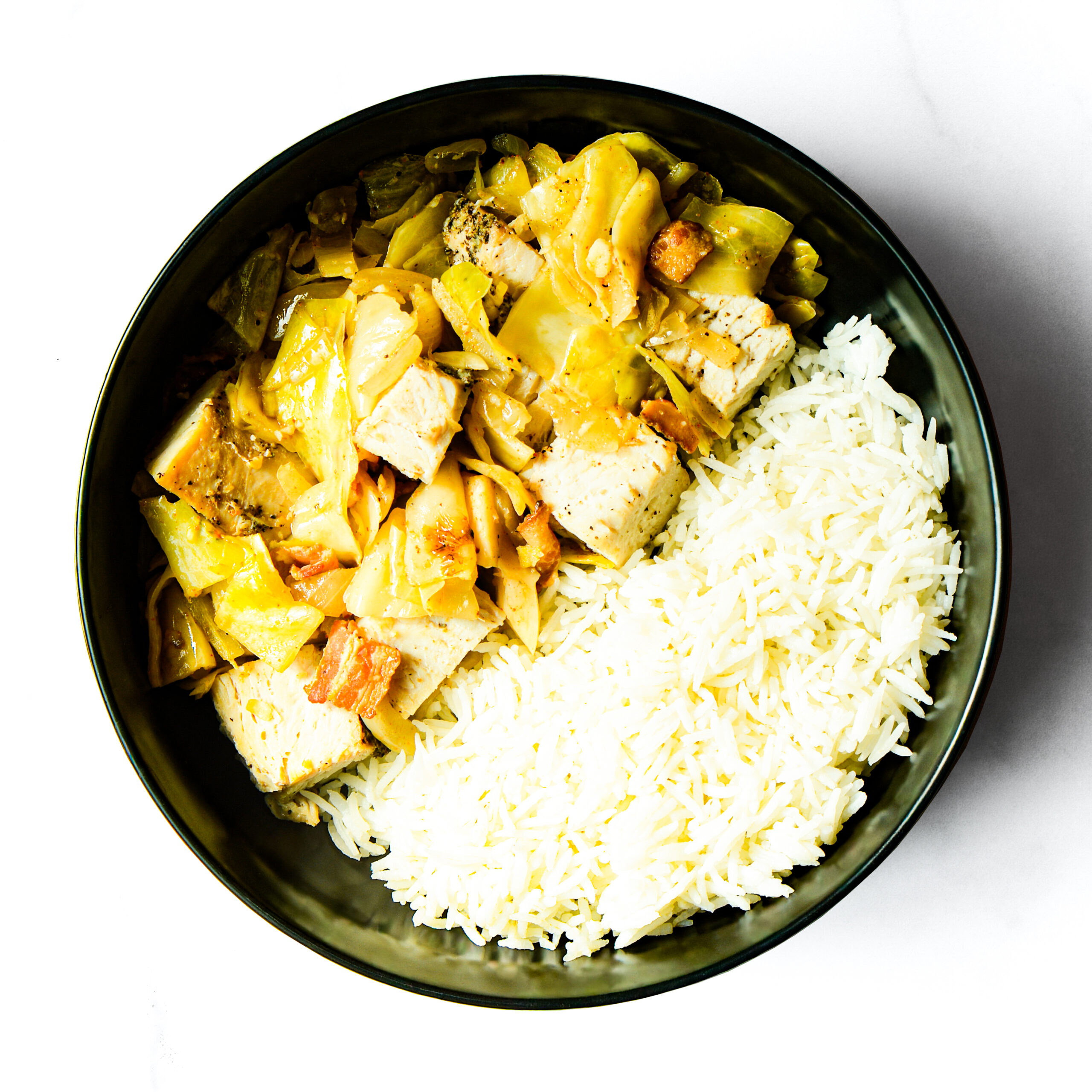 A black bowl of white rice topped with Operation Cajun Fit's cooked tofu, cabbage, and carrots sits on a white surface.