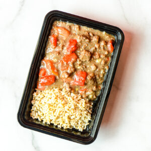 A black plastic tray of Mean Mexi-Machine, featuring cooked brown rice and a ground beef tomato stew, on a white surface.