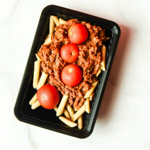A black plastic container of Southwest Shenanigans, You Clucker! with penne pasta, meat sauce, and four cherry tomatoes on a white surface.