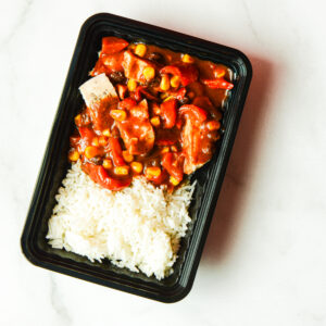 A black plastic tray of Southwest Shenanigans, You Clucker! with white rice and veggie stew in red sauce, on a white surface.