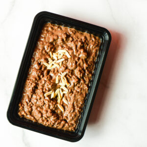 A black plastic container of Southwest Shenanigans, You Clucker!—a brown, chunky dish with slivered almonds—sits on a white surface.