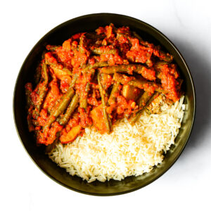 A black bowl containing Engine 38 Tortellini with white rice, green beans, and a red tomato-based vegetable stew.