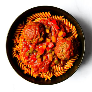 A black bowl of Engine 38 Tortellini topped with two meatballs and chunky tomato vegetable sauce on a white background.